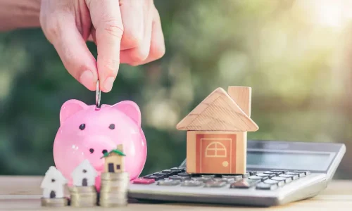 A hand is putting money into a piggy bank next to a house model.